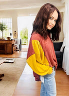 Woman wearing TRAVELING LIGHT top by BiBi with color block design in burgundy and gold, standing in a cozy living room.