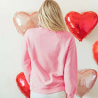 Back view of a woman in a pink LOVE YOU MORE CREWNECK against heart-shaped balloons.