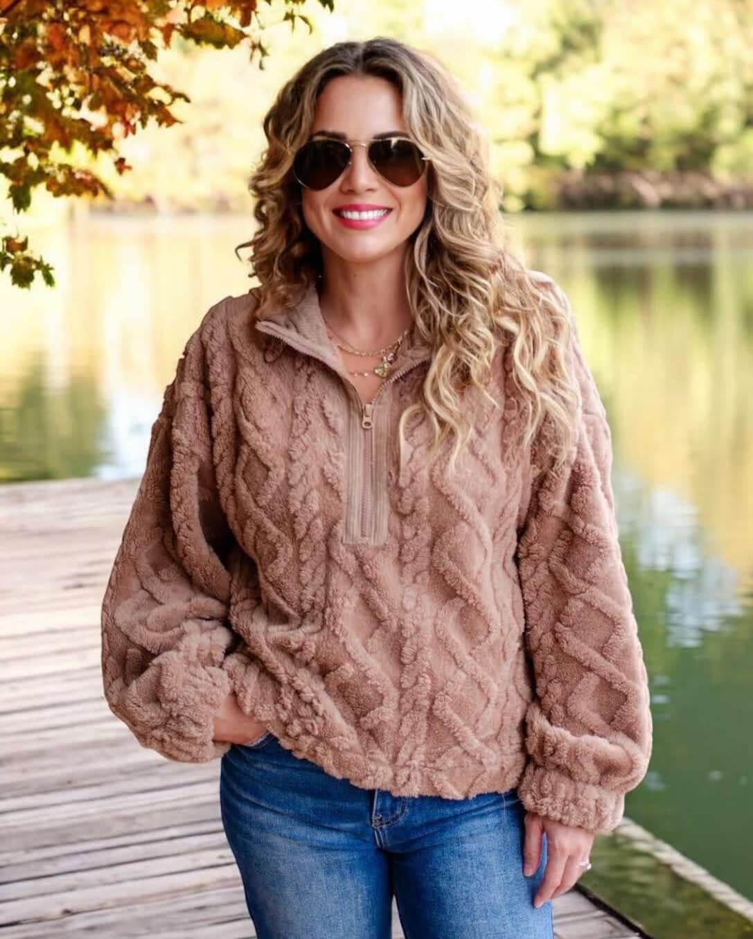 Woman wearing a mocha teddy bear sherpa fleece pullover with cable pattern design and half zip, standing outdoors by a lake.