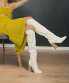 Woman wearing Stephanie White knee-high boots by Oasis Society, seated in a yellow dress showcasing high heel design.