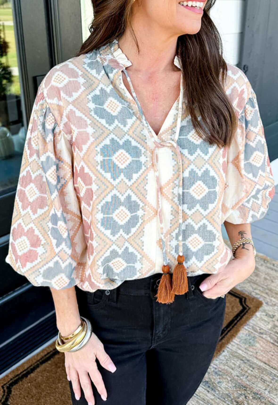 Woman wearing a trendy Painted Horizon Top with geometric designs and tassel details, styled with black pants.