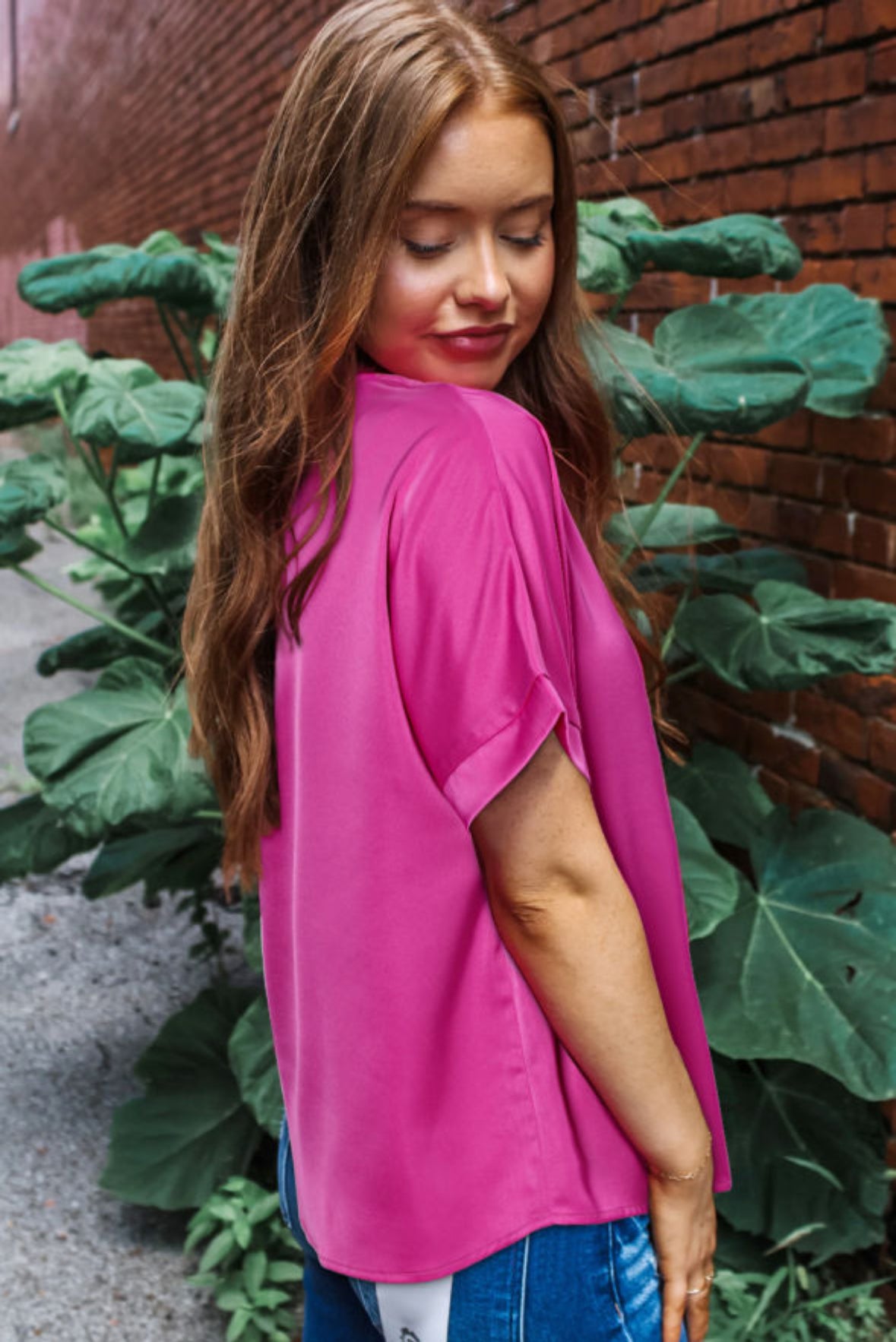 Stylish woman wearing a vibrant Raspberry Rush top, posing against a brick wall with green foliage.