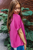 Stylish woman wearing a vibrant Raspberry Rush top, posing against a brick wall with green foliage.