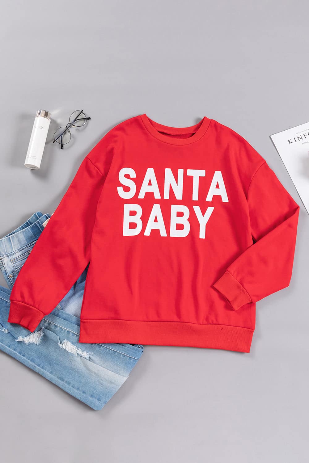 Red "Santa Baby" crewneck sweatshirt laid flat with light blue jeans and accessories for a festive holiday look.