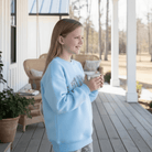 Girl wearing LULU MAC YOUTH SKY BLUE / CAMO CREW sweater holding a hot beverage on a porch.