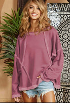 Stylish woman in a mauve relaxed fit top with exposed seams, paired with denim shorts in a trendy setting.