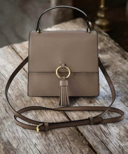 Taupe crossbody bag with gold ring and tassel detail, featuring faux vegan leather, detachable strap, and magnetic closure on a wooden table.