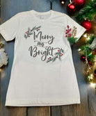 White Bella Canvas Merry and Bright t-shirt with holly trim, set among festive decorations on a wooden background.