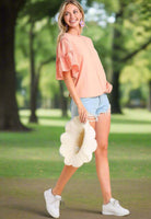 Model wearing Ruffled Breeze peach cotton top with ruffle sleeves and denim shorts in a sunny outdoor setting.