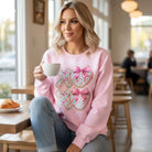 Model wearing LOVE YOU MORE CREWNECK in pink, featuring XOXO decal and bows, enjoying coffee in a cozy cafe.