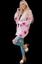 Woman wearing pink CARDI GIRL CARDIGAN with floral pattern, paired with ripped jeans and heels. Fashionable open-front knitwear.