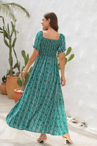 Model wearing a teal maxi dress with floral print, standing outdoors near cacti, showcasing the back and flowing design.