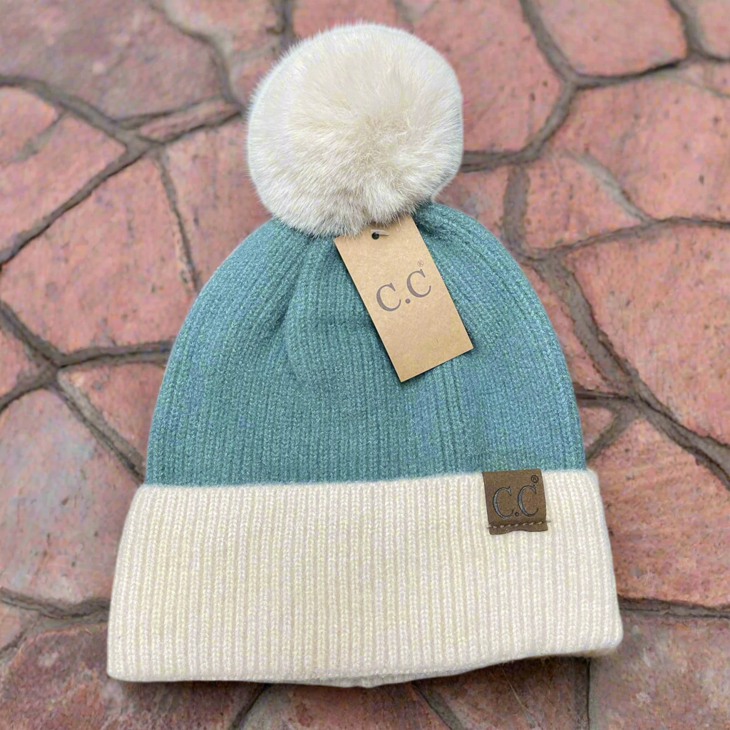 Teal OH SO SOFT BEANIE by C.C. with beige cuff and fur pom pom, featuring a suede patch, on a textured background.