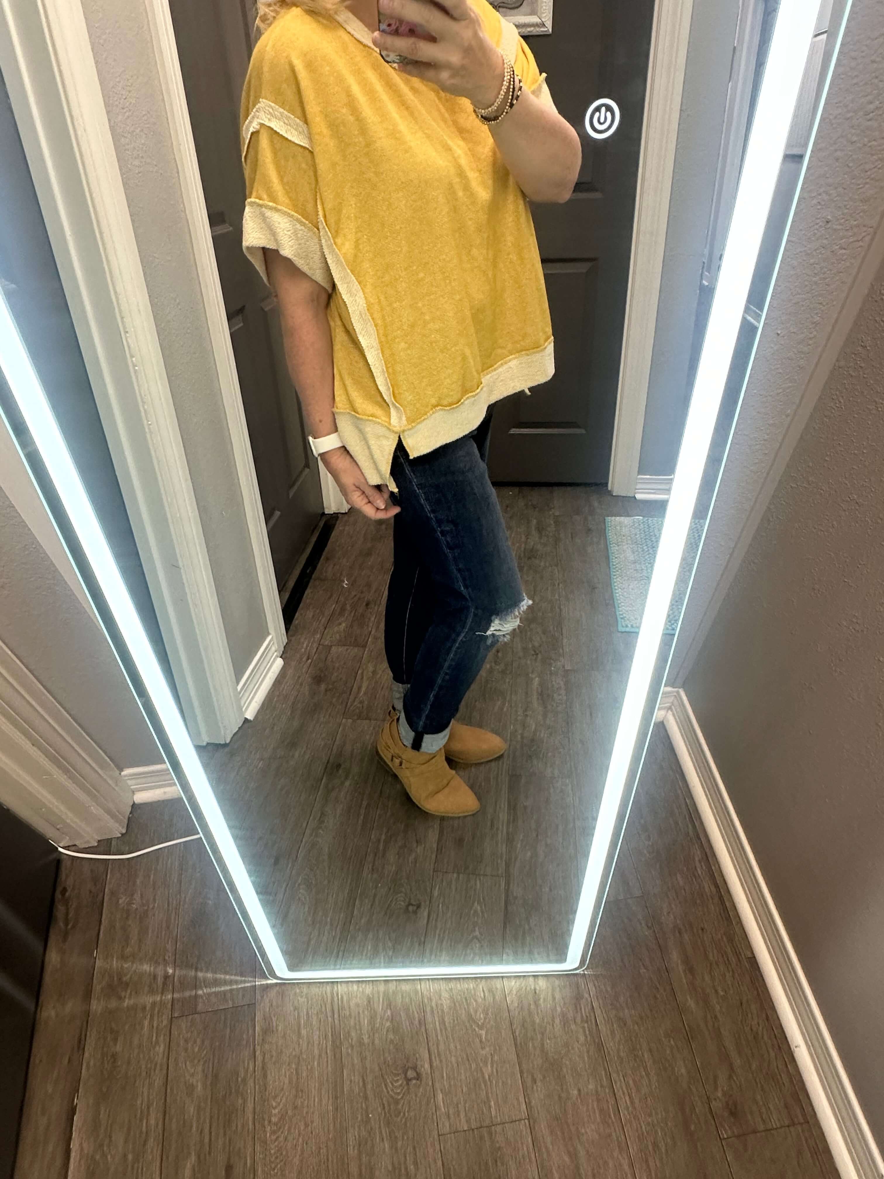 Woman in mellow yellow top with white accents and ripped jeans, posing in front of a full-length mirror.