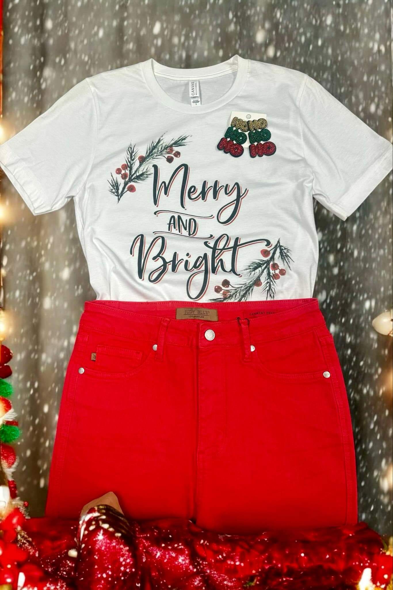 White Merry and Bright tee with holly trim by Bella Canvas, paired with red pants on festive background.