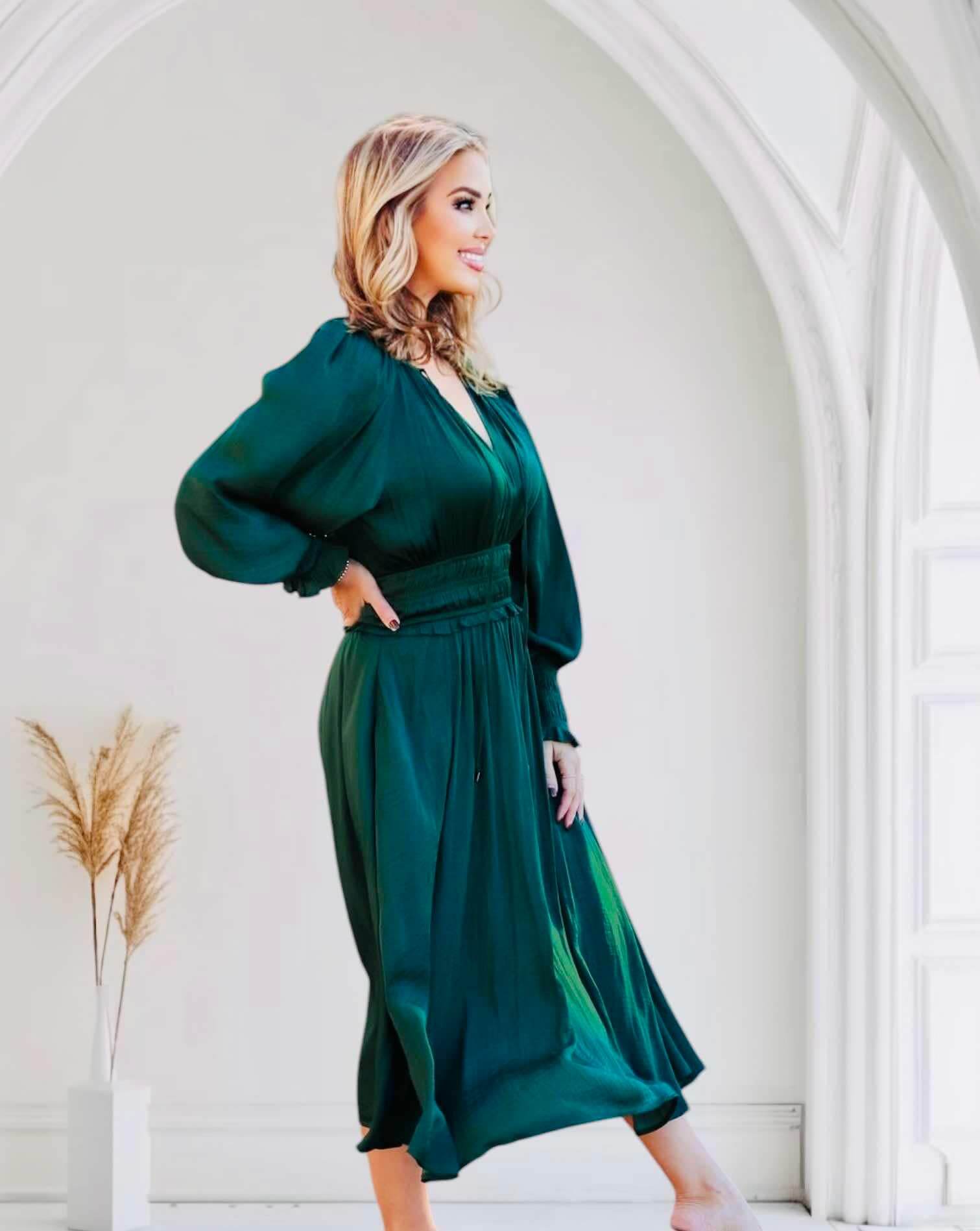 Woman wearing dark green midi dress with smocked sleeves and waist, standing in elegant arched room.