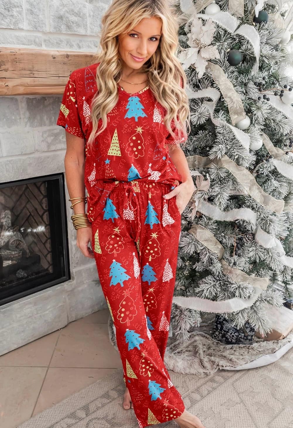 Woman wearing festive Christmas tree print pajamas near decorated holiday tree, cozy by the fireplace.