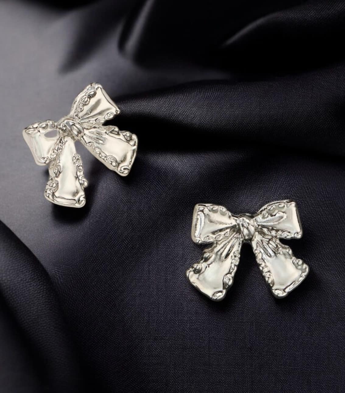 Silver bow stud earrings from the Vivian-Lu collection, showcasing an elegant design on a dark fabric background.