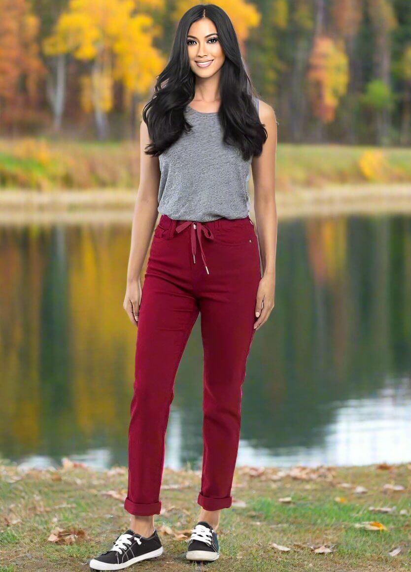 Woman wearing crimson Judy Blue Bridget Joggers with a drawstring waistband, standing outdoors by a lake in fall setting.