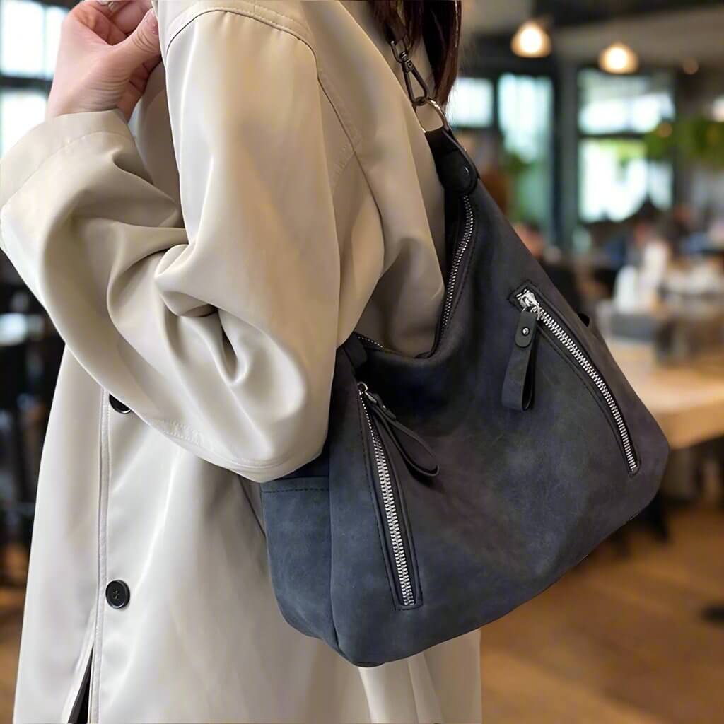 Woman wearing a beige coat and carrying a stylish faux suede Christina shoulder bag with zippers in a cozy indoor setting.