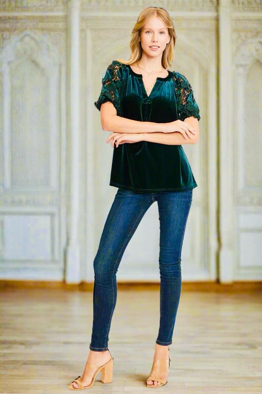 Woman wearing QUIET JOY Velvet Top in Dark Teal Green with lace sleeves, paired with jeans and heels, standing in an elegant room.