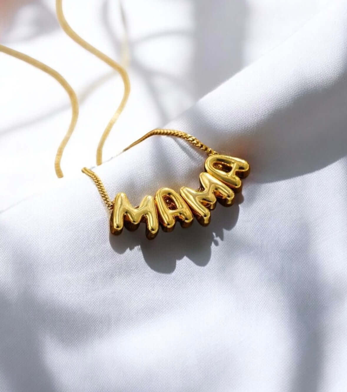 Gold tone Bubbly Mama Necklace with playful 'MAMA' charm on a light fabric background.