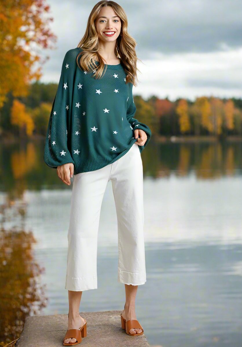 Woman wearing teal Umgee Seeing Stars sweater with star pattern, stylishly paired with white pants and brown sandals near a lake.