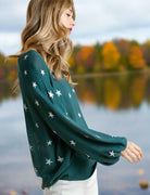 Woman wearing Umgee Seeing Stars Sweater in teal green with star pattern, standing by a lake with autumn foliage background.