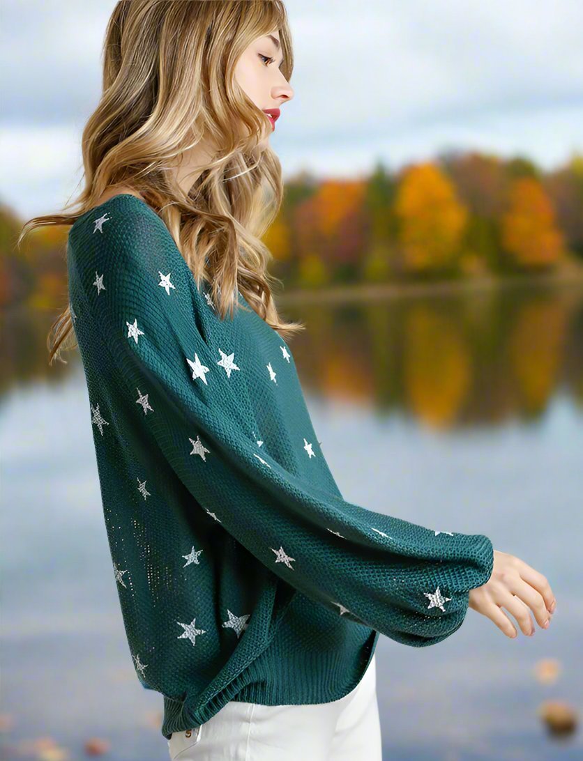 Woman wearing Umgee Seeing Stars Sweater in teal green with star pattern, standing by a lake with autumn foliage background.