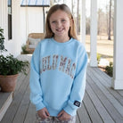 Young girl wearing LULU MAC YOUTH SKY BLUE / CAMO CREW sweatshirt, smiling on a porch.