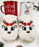 White reindeer-themed slippers with red bow and antlers, paired with holiday-themed mugs labeled "warm & cozy," perfect for winter.