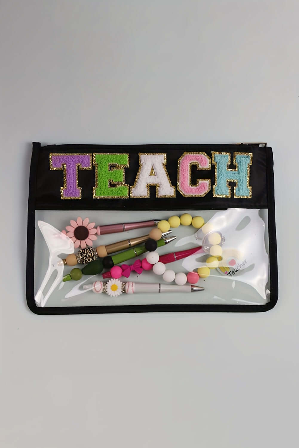 Clear zipper pouch with 'TEACH' embroidered, showcasing colorful pens and accessories inside.