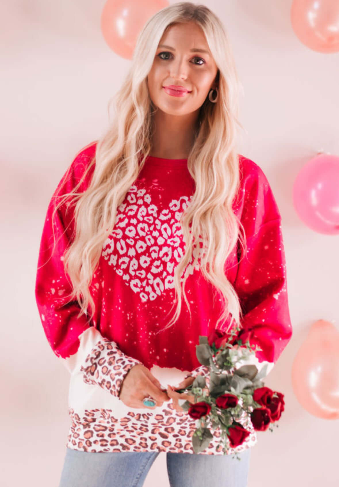 Model wearing Wild Hearts Crewneck with leopard heart design, holding red roses against a backdrop of pink balloons.