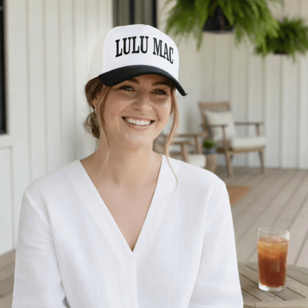 A woman wearing a LULU MAC BLACK HAT, smiling outdoors with a drink beside her, showcasing stylish casual wear.