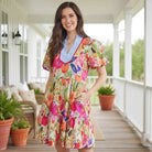 Model wearing BRIGHT DAYS AHEAD DRESS featuring floral print, striped patchwork, and ricrac trim on a porch.