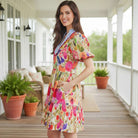 Model wearing BRIGHT DAYS AHEAD DRESS with floral print and striped trim, posing on a porch surrounded by greenery.
