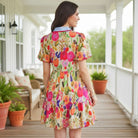 Model wearing the BRIGHT DAYS AHEAD DRESS featuring vibrant floral patterns and ricrac trim, set on a porch.