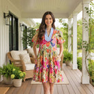 Model wearing BRIGHT DAYS AHEAD DRESS with floral print and ricrac trim, posing on a porch surrounded by greenery.