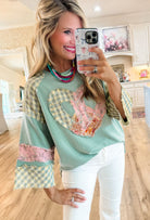 Woman wearing mint PEACEKEEPER top with plaid and floral patches, peace sign and heart graphic, wide sleeves, in a casual setting.