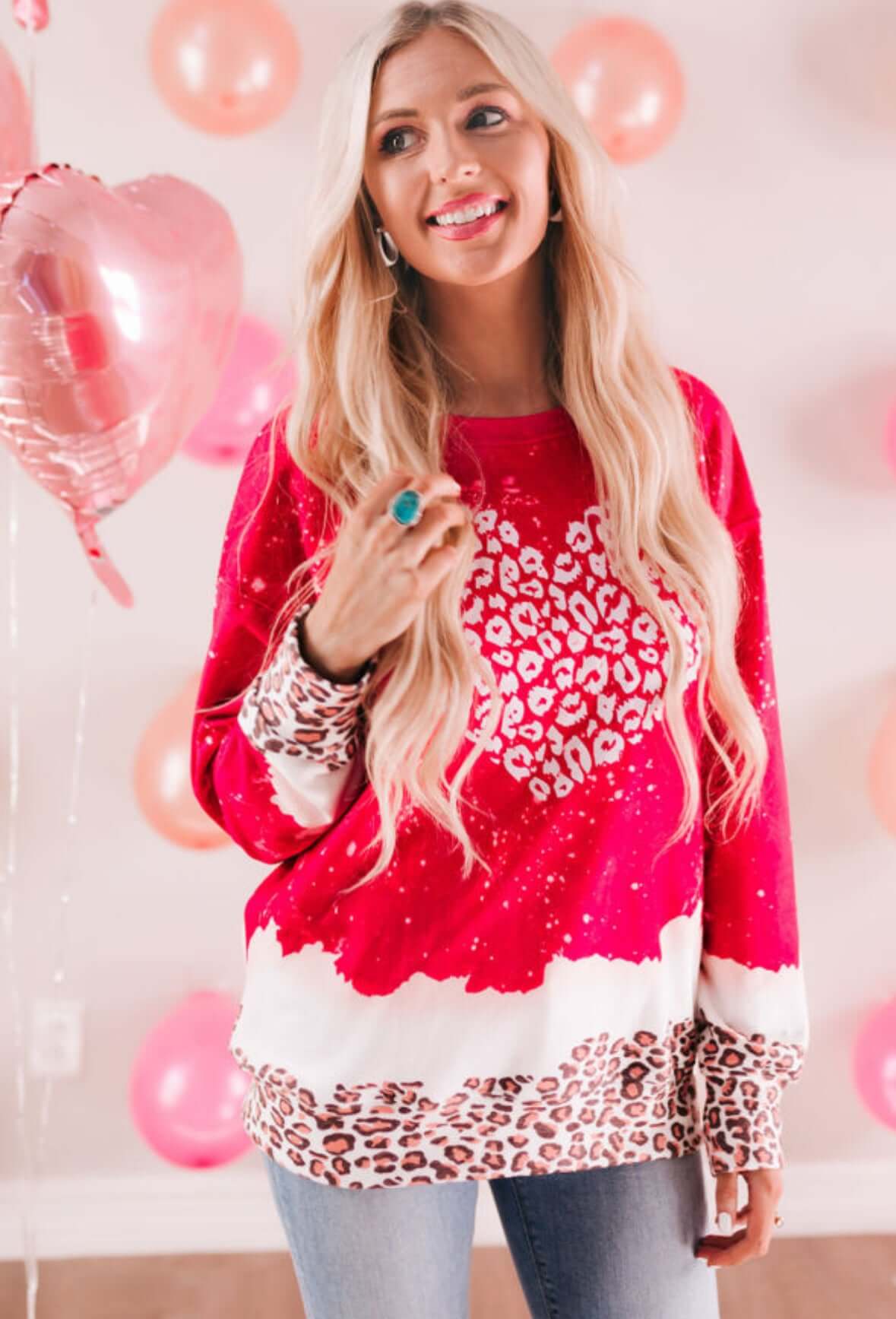 Young woman in a pink and leopard print Wild Hearts Crewneck sweater, smiling with heart-shaped balloons in the background.