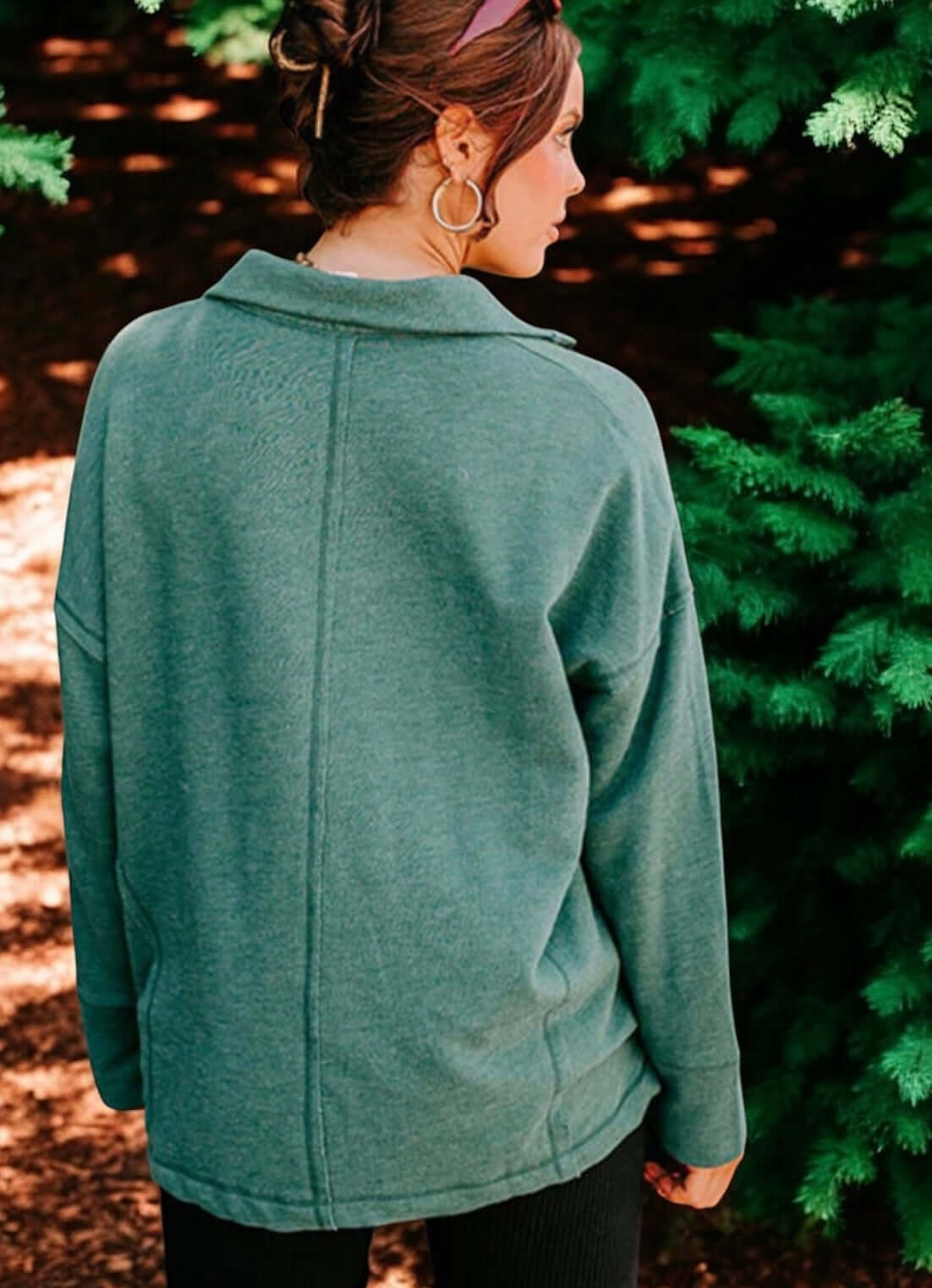 Woman wearing an evergreen exposure oversized sweater, standing amid lush green trees, showcasing a relaxed back view.