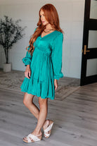 Woman wearing teal green Bimini Beauty Dress with v-neckline and balloon sleeves, standing in a stylish indoor setting.