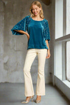 Woman wearing teal velvet top with pearl-embellished lantern sleeves and high-low hem, standing by a window.