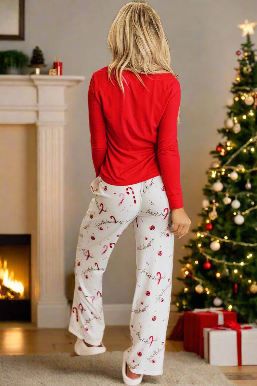 Woman wearing Sweet Dreams Candy Cane pajamas in front of a Christmas tree and fireplace, featuring red top and candy cane pants.