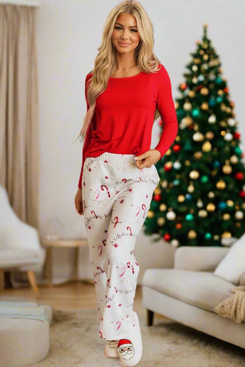 Woman wearing Sweet Dreams Candy Cane pajamas with red top and festive pants, standing in a cozy holiday living room with decorated tree.