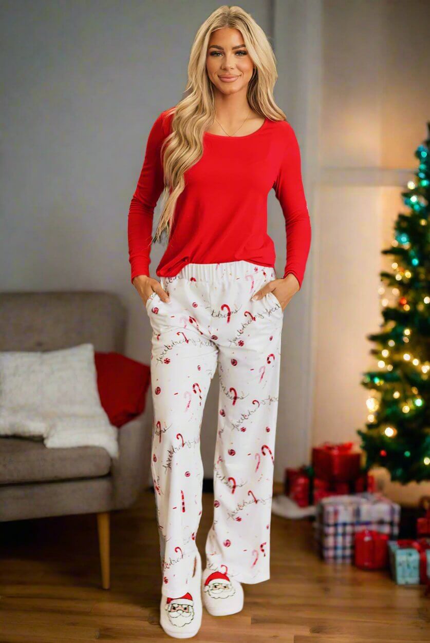 Woman wearing SWEET DREAMS CANDY CANE pajamas with red top and candy cane print pants, standing near Christmas tree and gifts.