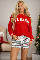 Woman wearing SLEIGH GIRL SLEIGH PJ Set with red top and snowflake print shorts, standing by a Christmas tree.