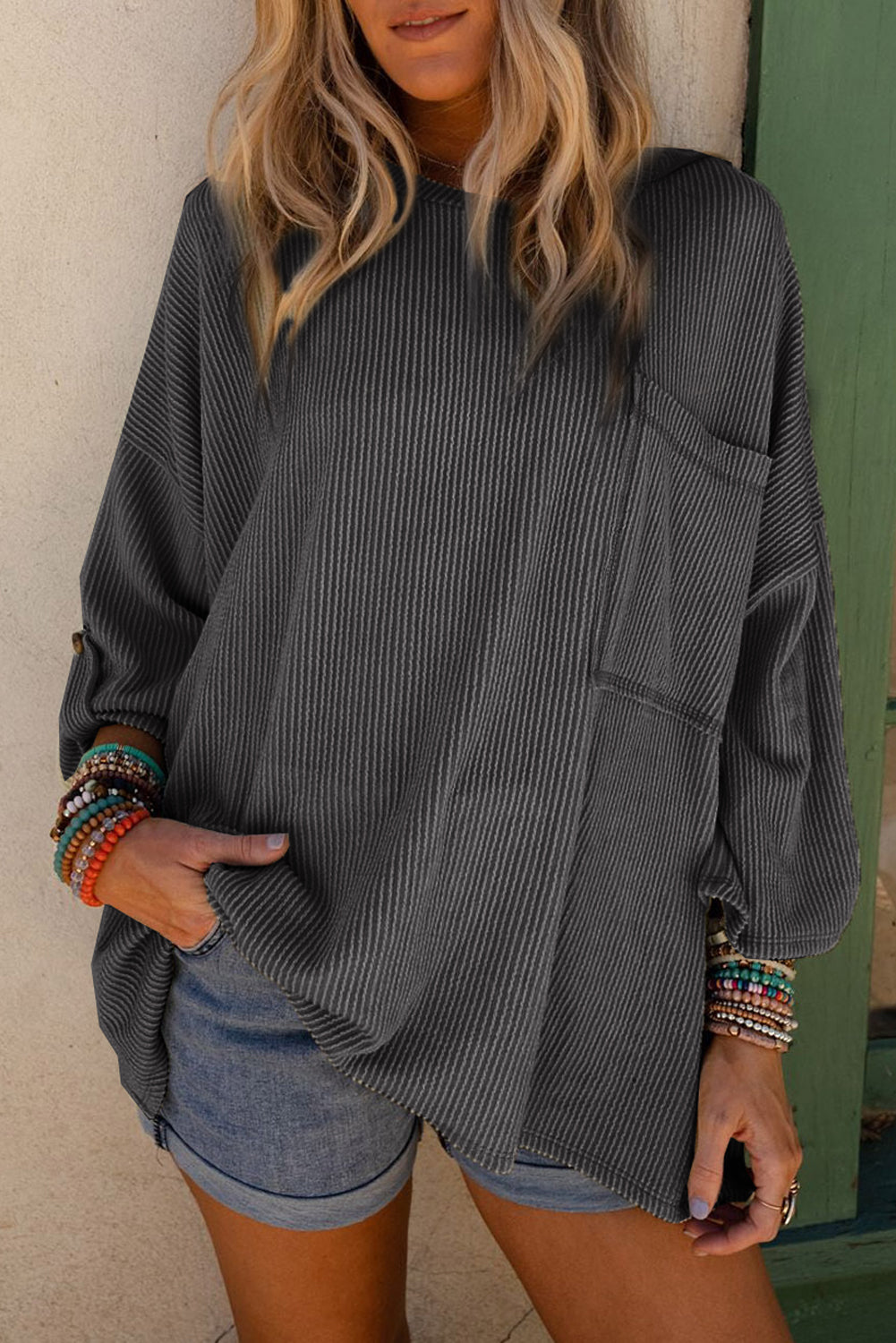 Woman wearing oversized charcoal ribbed knit top with roll-tab sleeves, paired with denim shorts and colorful bracelets.