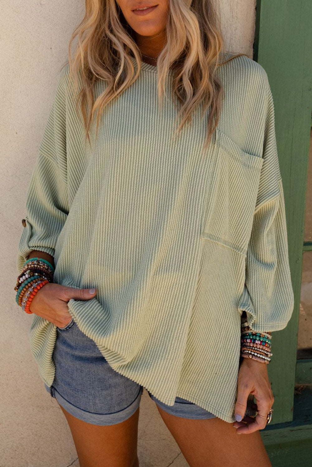 Woman wearing Lines Drawn oversized sage top with ribbed knit and roll-tab sleeves, paired with denim shorts.