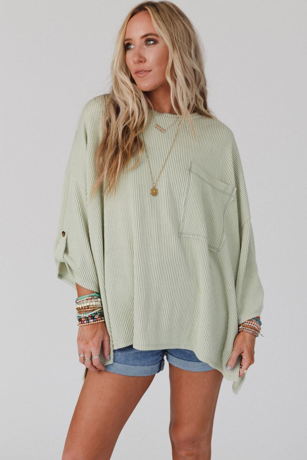 Woman wearing sage Lines Drawn Top, oversized with roll-tab sleeves and chest pocket, styled with denim shorts and layered bracelets.